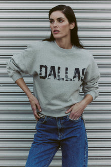 women wearing a gray sweatshirt with DALLAS printed on it standing in front of a metallic shutter