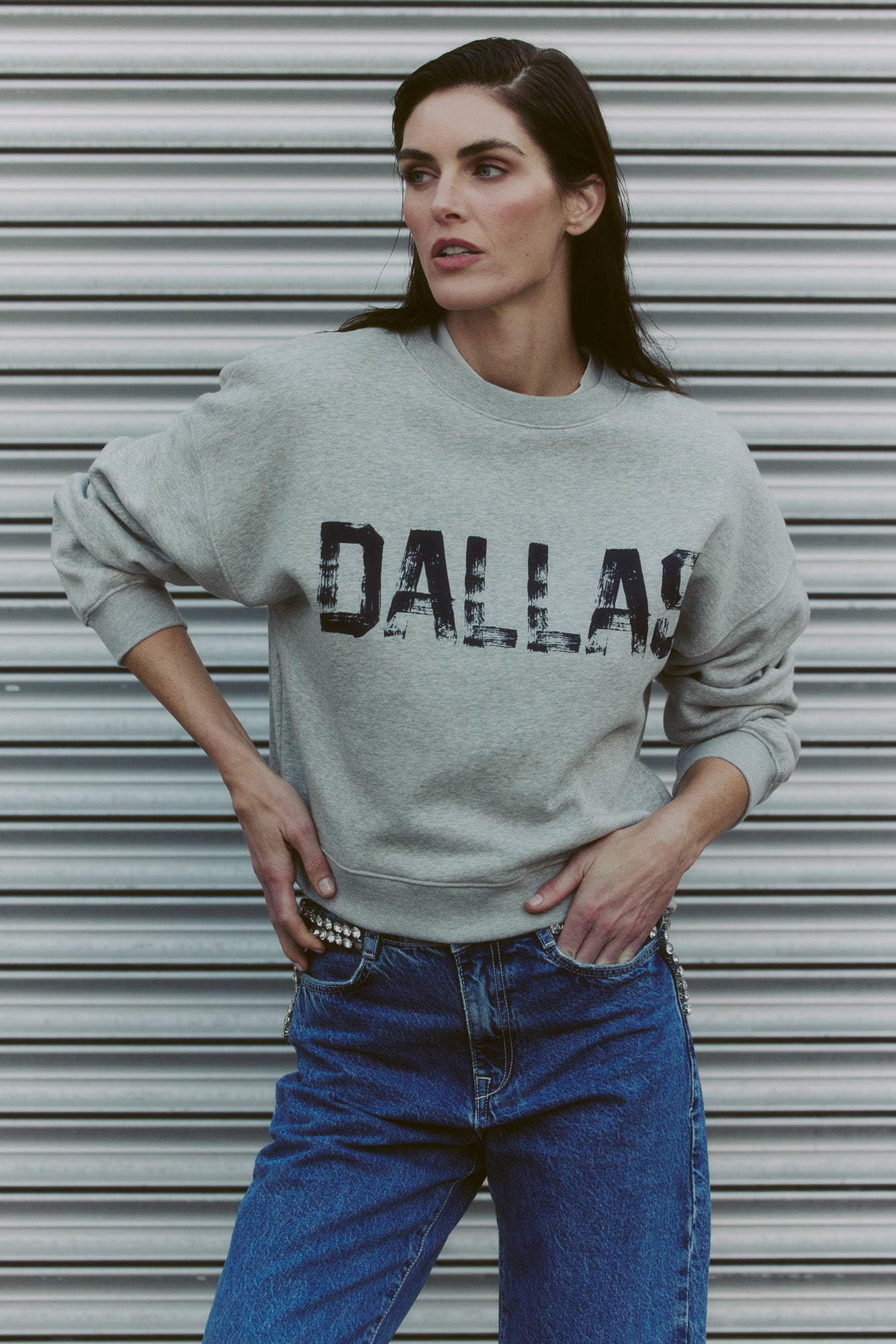 women wearing a gray sweatshirt with DALLAS printed on it standing in front of a metallic shutter