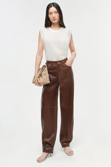 Woman wearing a cream sleeveless cashmere vest with brown leather pants and holding a beige bag.
