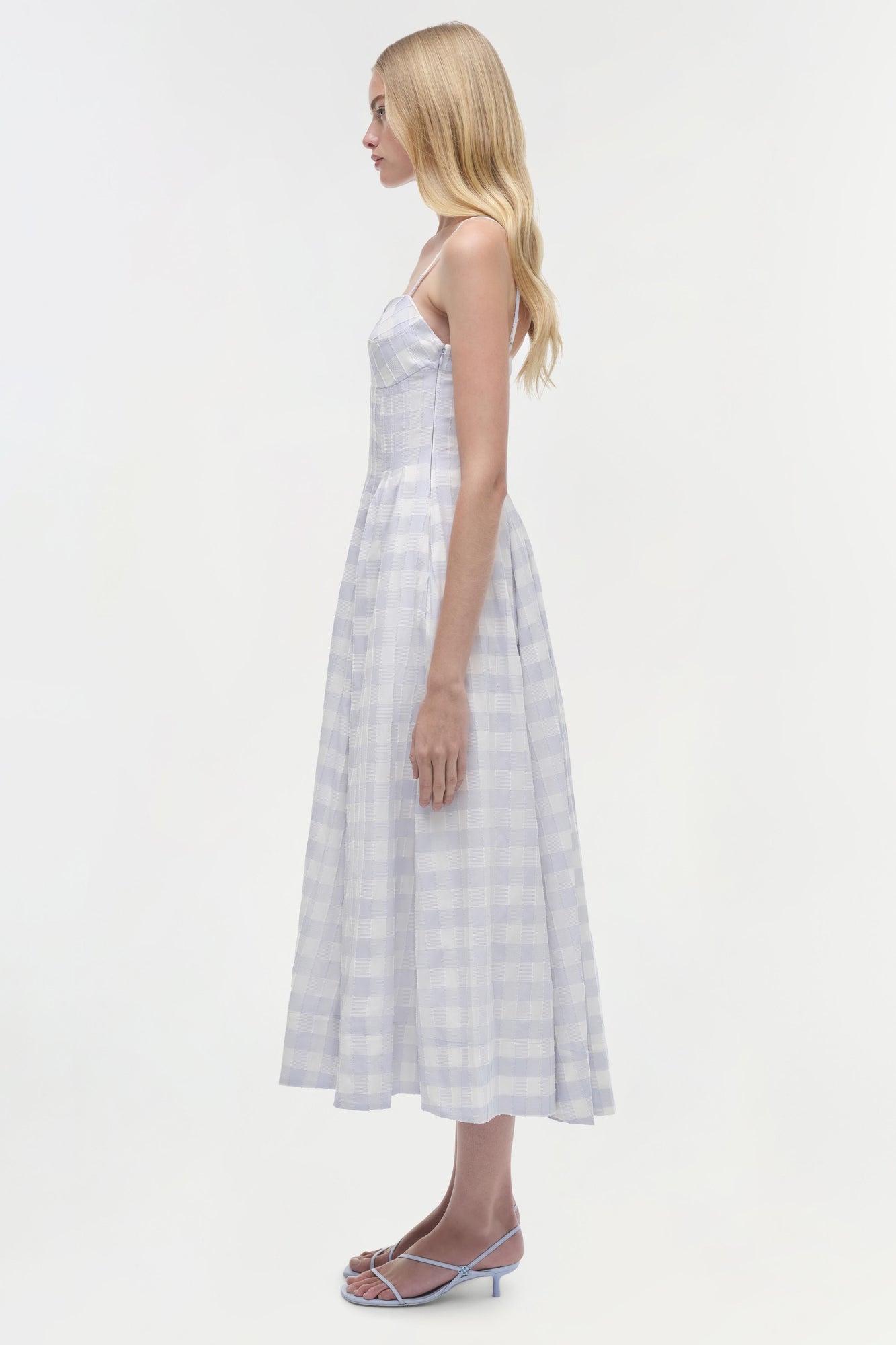 Woman wearing light blue and white gingham midi dress with thin straps, shown from the side.