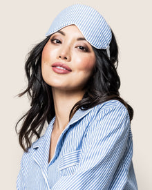An individual with long dark hair wears Petite Plumes Adults Sleep Mask in French Blue Seersucker atop their head, complemented by matching blue and white striped pajamas. Their neutral expression exudes luxury sleep against a light background.