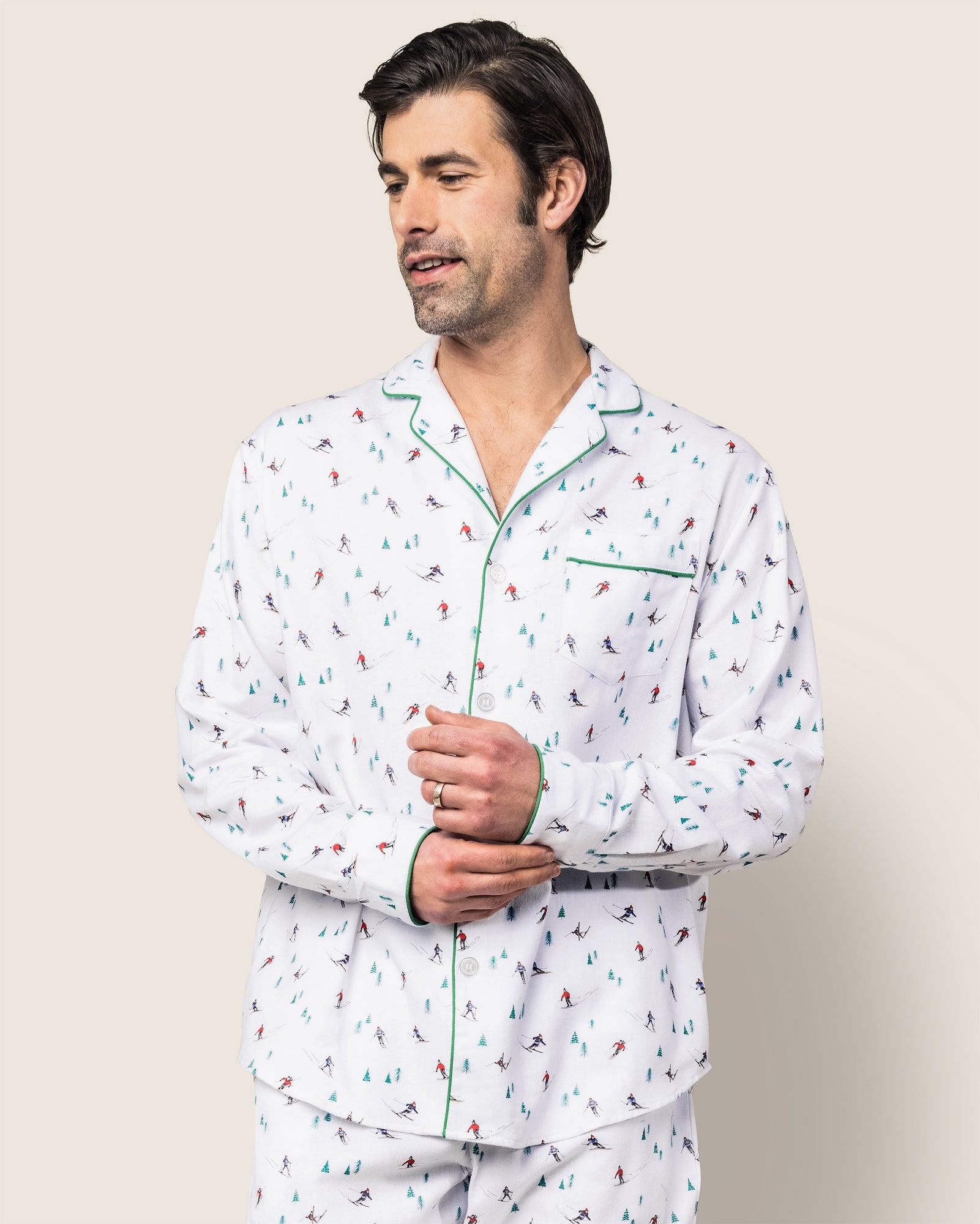 A man models the Mens Flannel Pajama Set in Apres Ski by Petite Plume. The classic white sleepwear showcases colorful skier prints and features green piping along the collar and pocket, adding a timeless touch to this luxury set against a plain background.