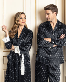 A woman with long wavy hair and a man with short hair stand side by side, wearing Petite Plumes matching black Art Nouveau silk pajamas. They are in a room with cream-colored paneled walls.