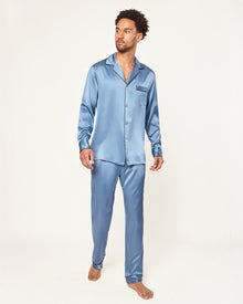 The Men's Silk Pajama Set in Mystique Blue features a button-up long-sleeve top with a chest pocket and matching elastic-waist pants, both trimmed with dark piping.