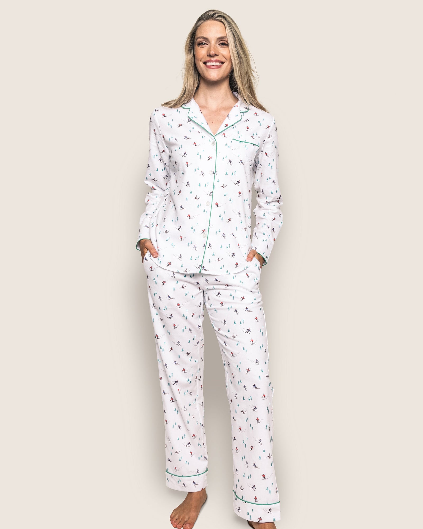A person stands smiling, wearing the luxurious Womens Flannel Pajama Set in Apres Ski by Petite Plume. The top features green piping and a collar, while hands are in the pockets of loose-fitting, soft brushed cotton flannel pants. The background is a plain light color.