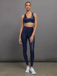Model wears a navy, glossy, deep scoop-neck sports bra with a sleek racerback and a shiny, liquid-like finish.
