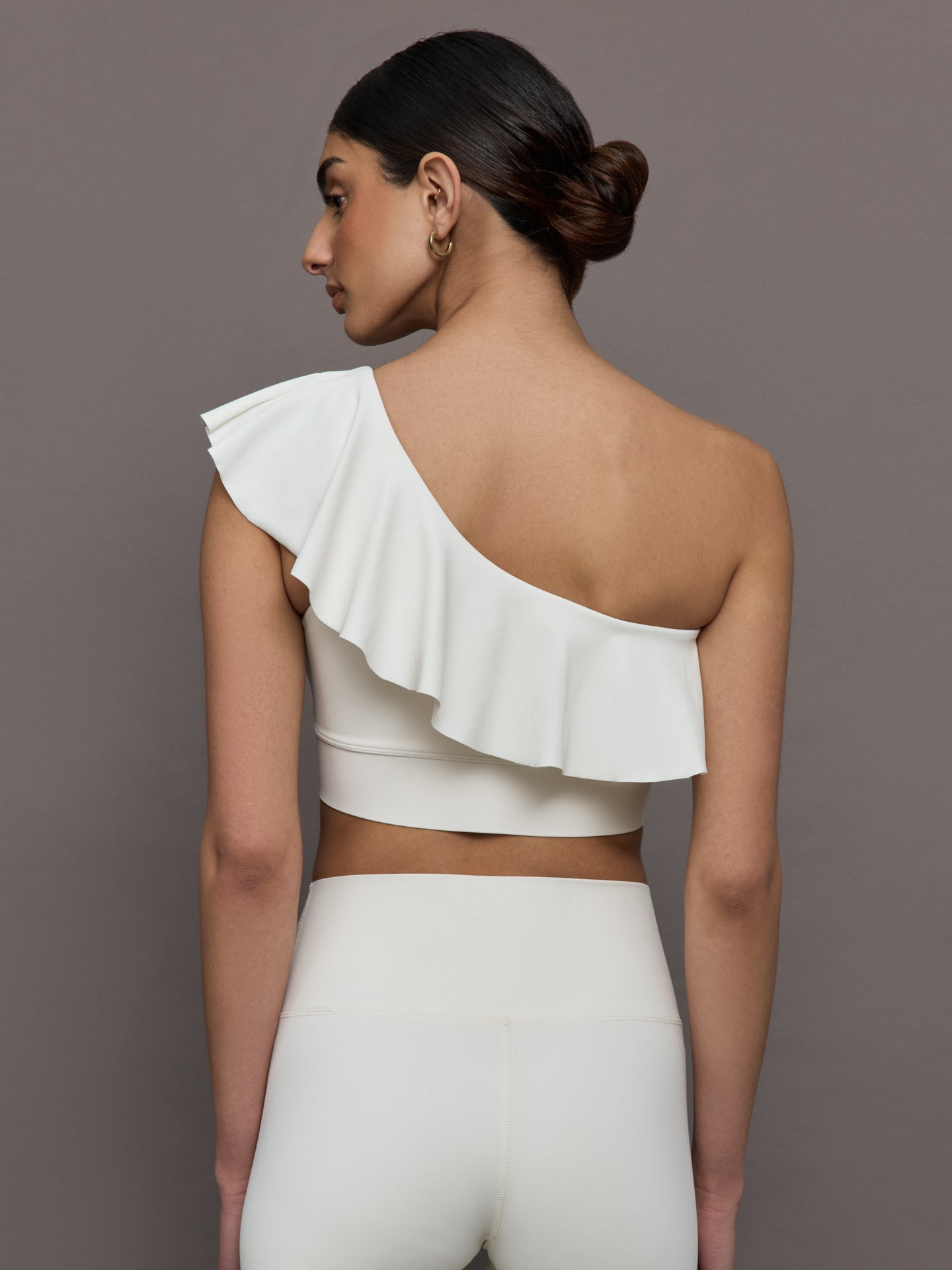 Model wears a white one shoulder sports bra with a ruffled overlay. 