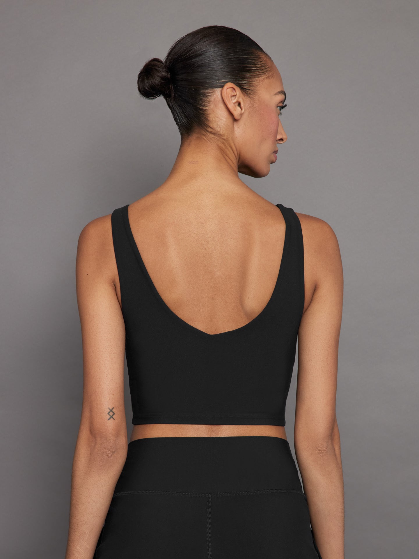 Model wears a black, longline sports bra with a deep V-neck front and an open V-shaped back, made from sleek, smooth fabric.
