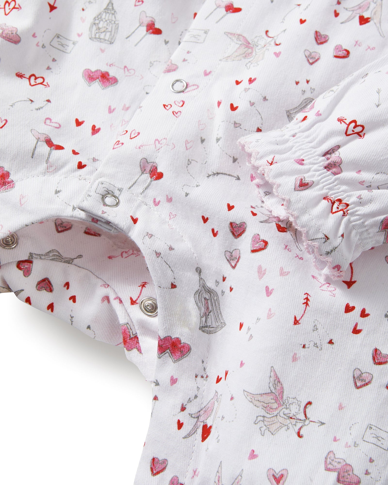 The Baby's Twill Madeline Romper in Love Song is a white long-sleeve romper with ruffled cuffs and collar, decorated with a pink pattern of hearts, glasses, and small animals. It features snap buttons down the front and legs.