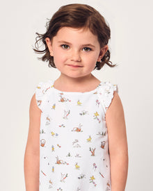 A young girl with short brown hair smiles in the Girl's Twill Amelie Nightgown in Easter Frolic, a white sleeveless dress featuring playful animal and nature prints, standing against a plain light background.