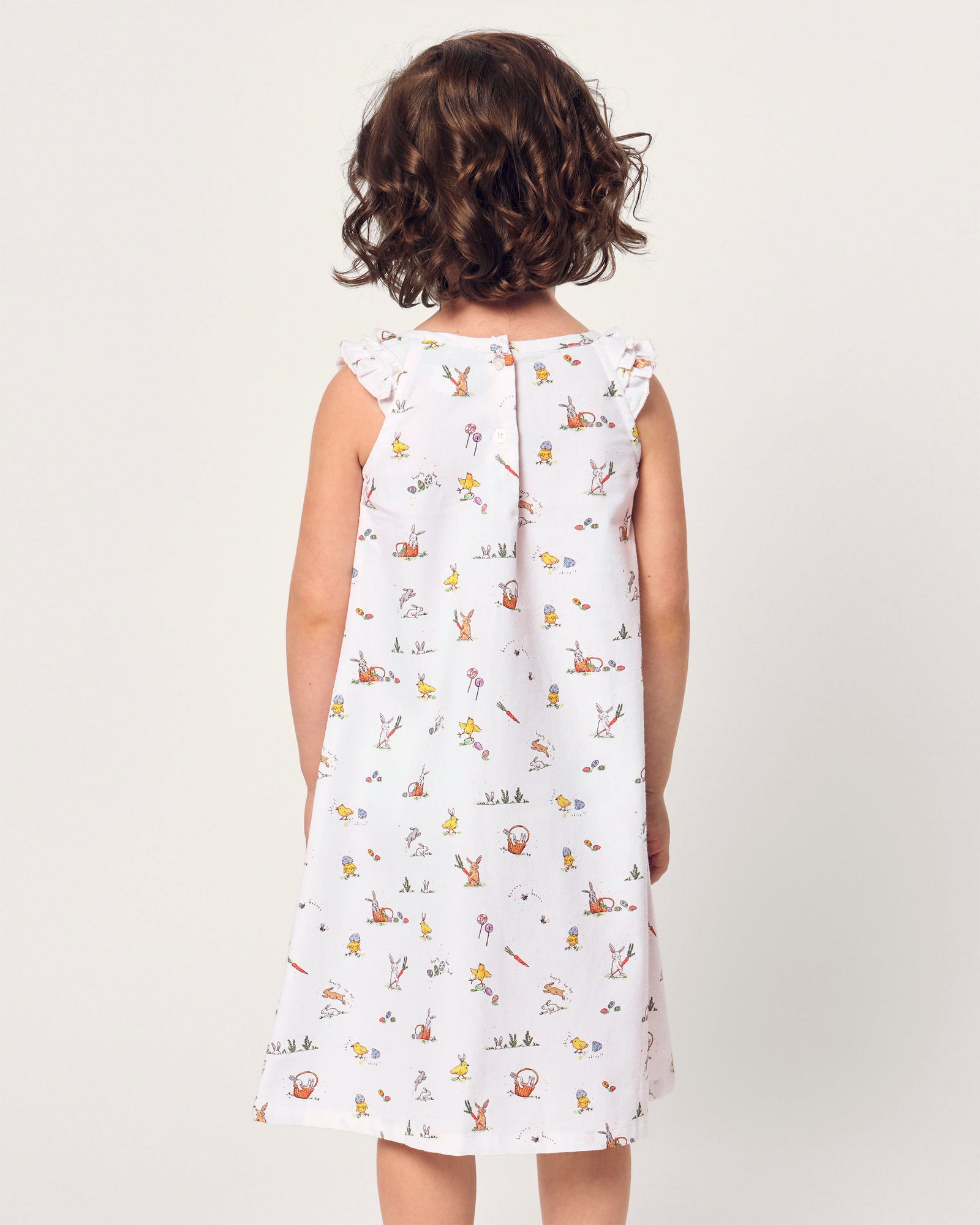 A young girl with short brown hair smiles in the Girl's Twill Amelie Nightgown in Easter Frolic, a white sleeveless dress featuring playful animal and nature prints, standing against a plain light background.