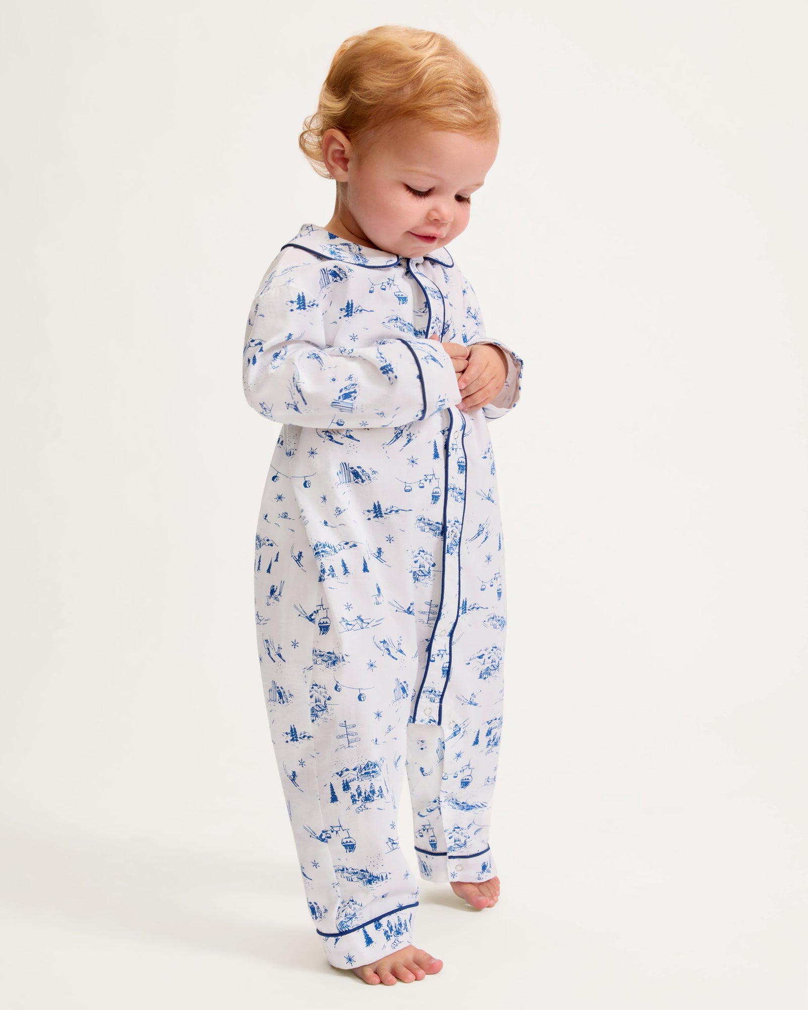 The Baby's Twill Cambridge Romper in Chalet Toile is a white long-sleeve, full-length onesie with blue alpine scene prints and blue piping on the collar, cuffs, and front placket.