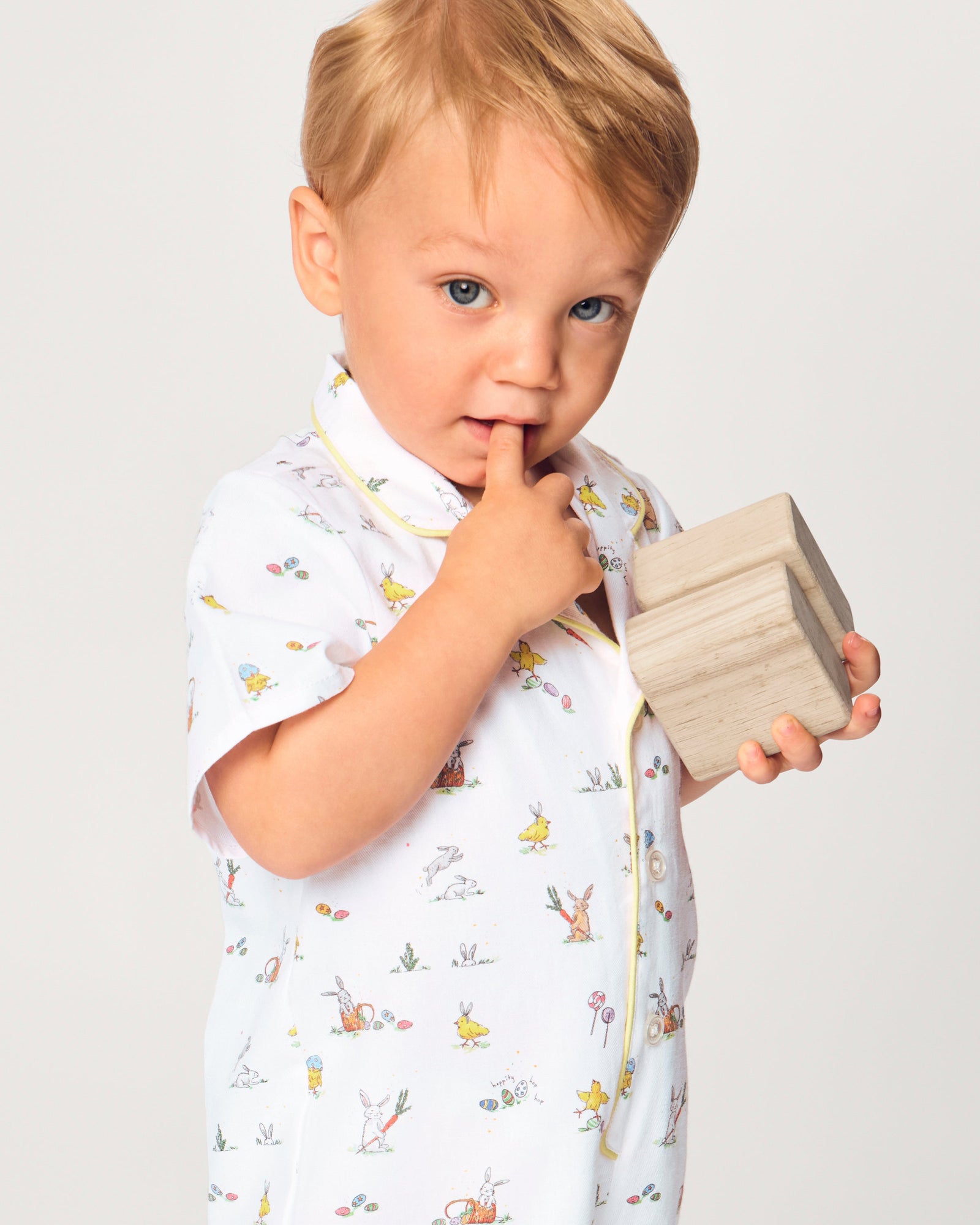 A young child with blonde hair wears the Baby's Twill Summer Romper in Easter Frolic—white with colorful animal prints and yellow trim—standing against a plain light background, holding two wooden blocks and looking to the side.