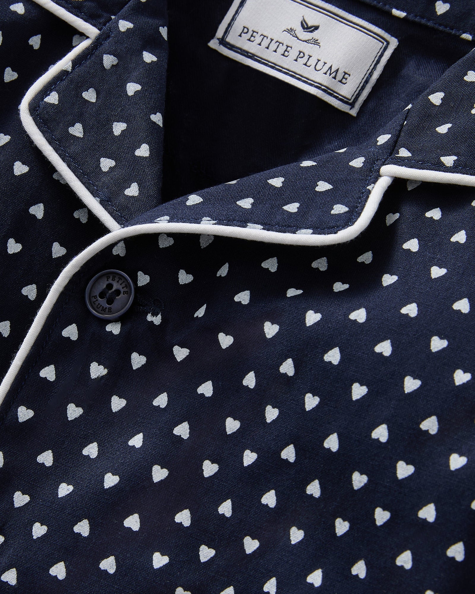 The Baby's Twill Classic Romper in Midnight Hearts is a navy blue onesie with long sleeves and legs, featuring a white heart pattern and white piping on the collar, cuffs, and front.