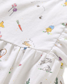 A young child with brown hair in pigtails wears the Baby's Twill Ruffled Romper in Easter Frolic, featuring colorful animal and floral prints, standing against a plain light background.