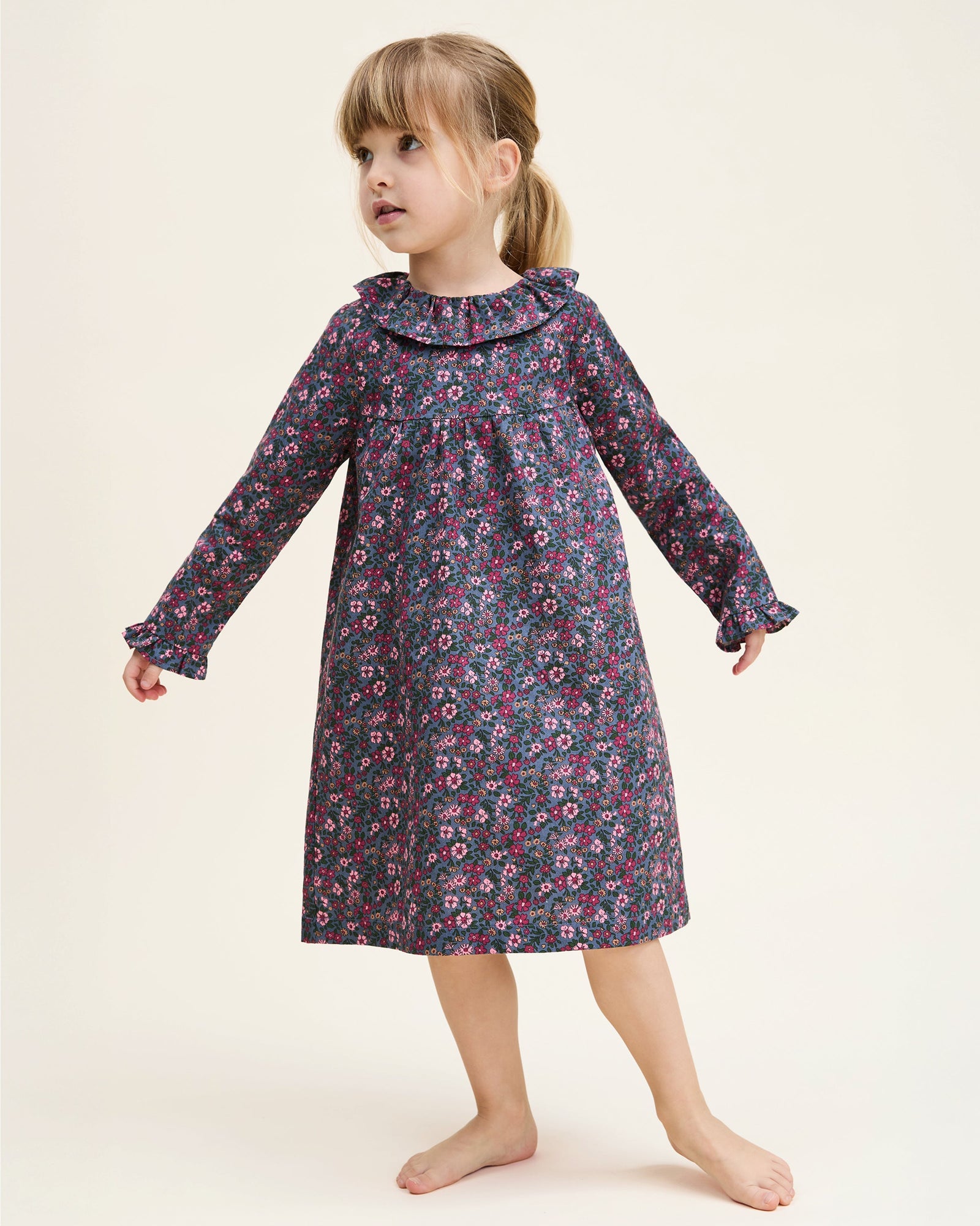 The Girl's Twill Scarlett Nightgown in Fleurs de Saison is a long-sleeved, knee-length dress with a ruffled collar and cuffs, featuring pink and white flowers with green leaves on a dark background.