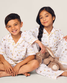 Two smiling children in luxurious Petite Plume sleepwear sit on the floor. They sport matching Kids Twill Short Sets in an Easter Frolic print. One child cuddles a stuffed bunny, embodying comfort and joy against a plain backdrop.