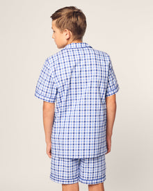 A young boy faces away, dressed in Petite Plumes Kids Twill Pajama Short Set in Royal Blue Gingham, made of yarn-dyed cotton with short sleeves and matching shorts. The background is plain off-white.
