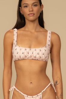 Pale pink bikini top with cherry print and thick padded straps on model against neutral background.