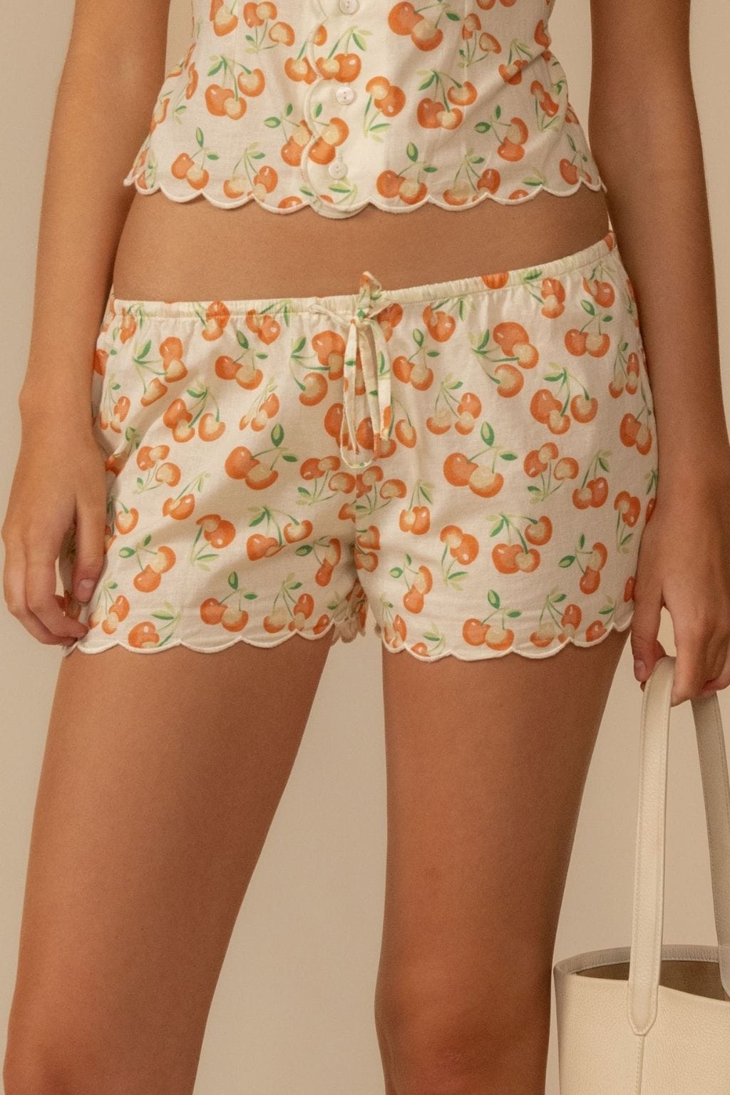 Cream shorts with orange cherry and green leaf print, scalloped edges, and drawstring waist.