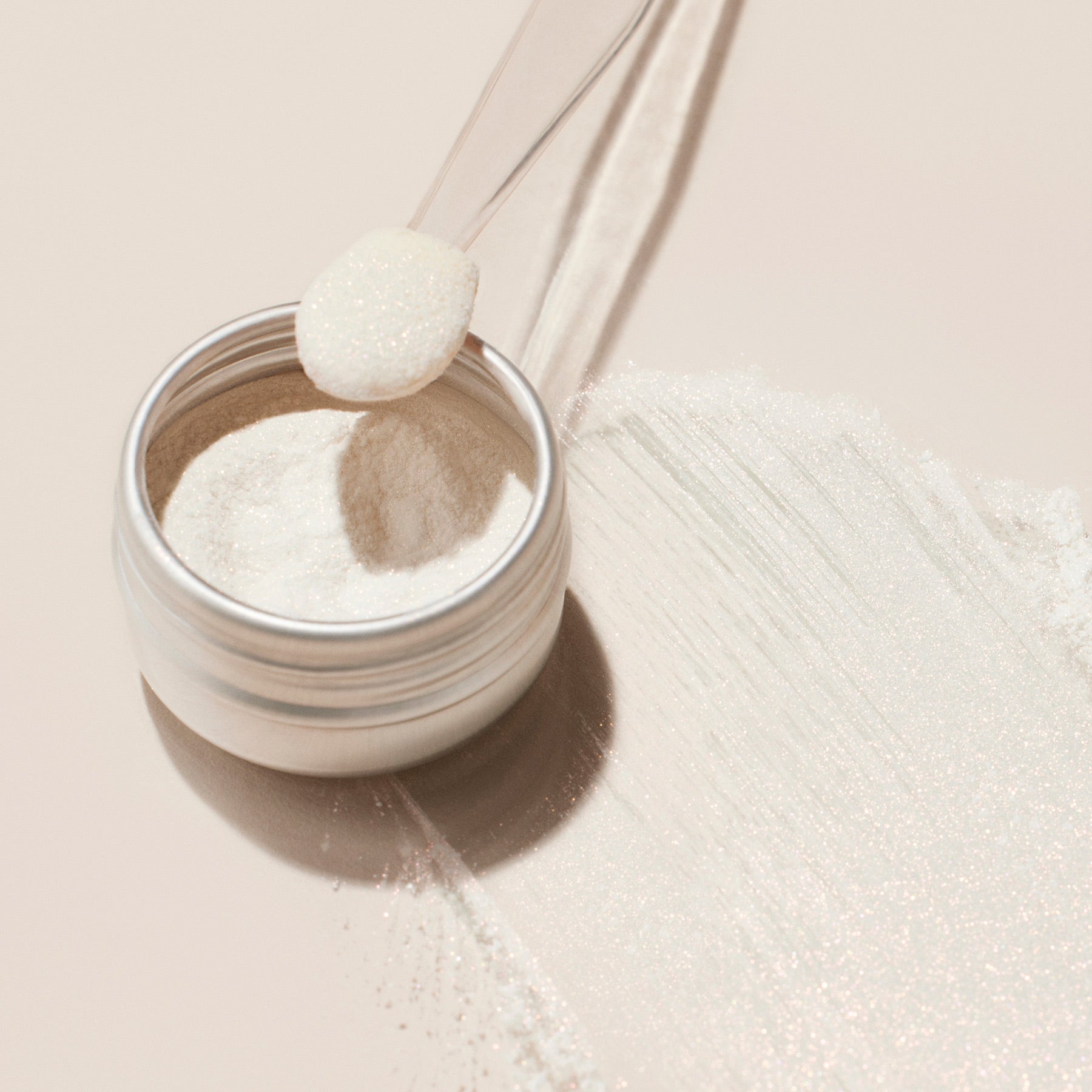 A small metal jar of Manucurist's Glitters Duo Rose Gold  Glazed sits on a light surface. A plastic scoop holds some pearlescent powder, with more of the sparkling product spilled and streaked nearby.