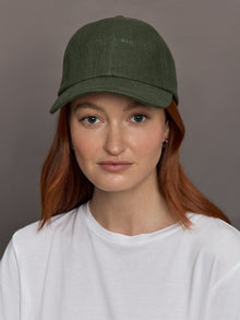 Model wears olive linen baseball cap with an embroidered logo on the front and a ruched elastic back.