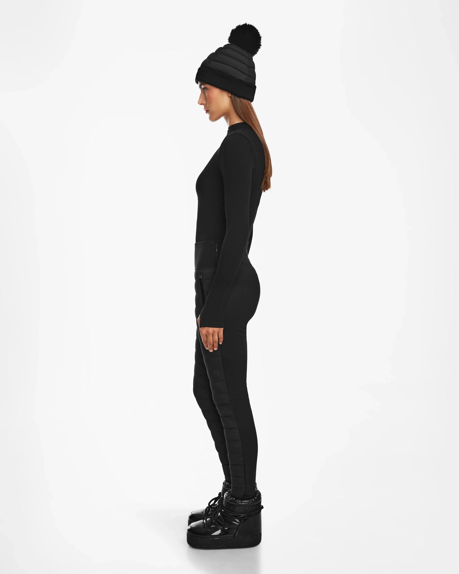 Woman in side profile wears Rudsak ARIL M - 8522514 BLACK beanie with pom-pom, a black long-sleeve top, après-ski chic womens quilted pants, and black platform boots, standing against a white background.