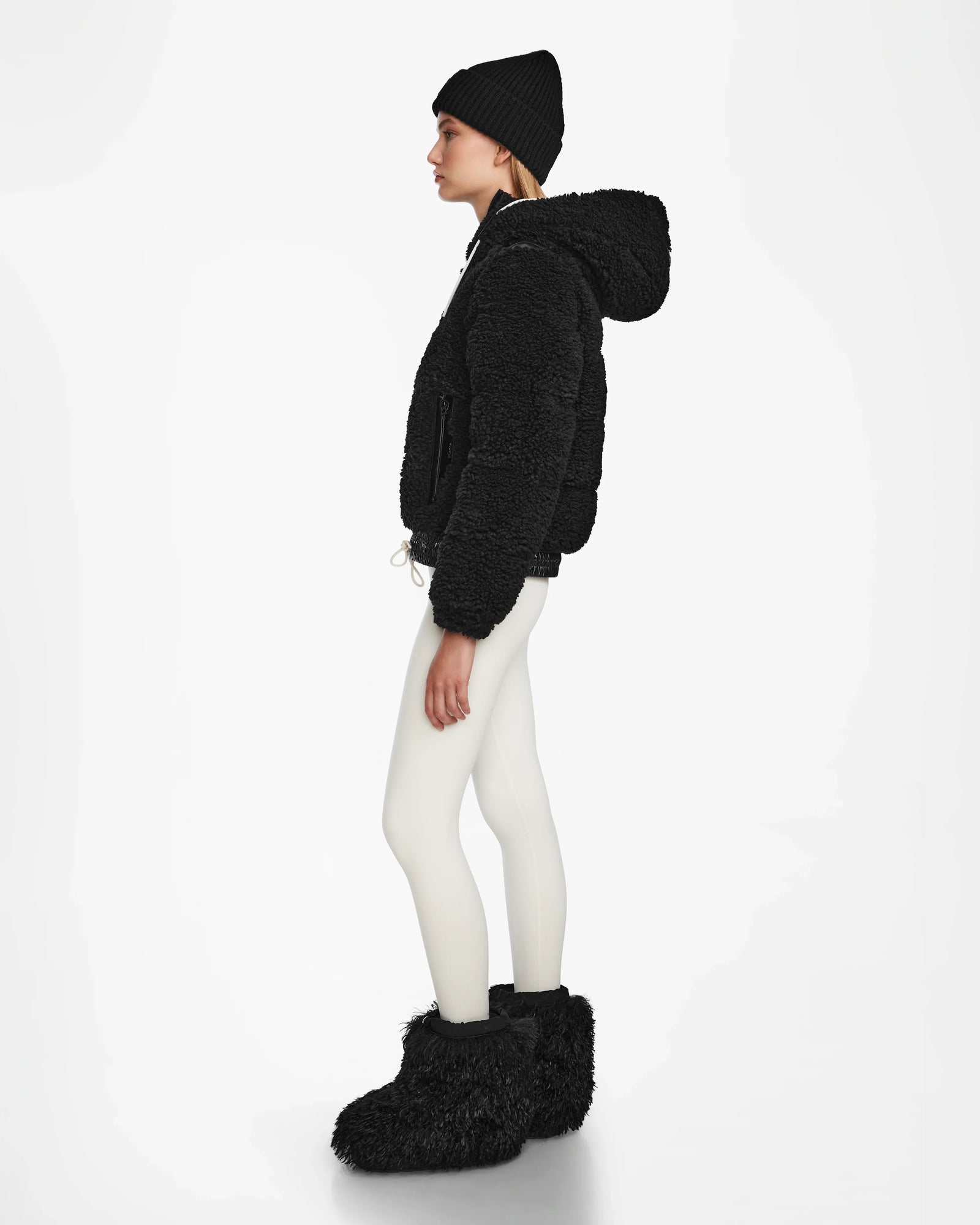 A woman stands in profile against a white background, wearing the Rudsak DANI S - 8124537 Black cropped ski jacket with a fuzzy hood, paired with a black beanie, white leggings, and large black fuzzy boots.