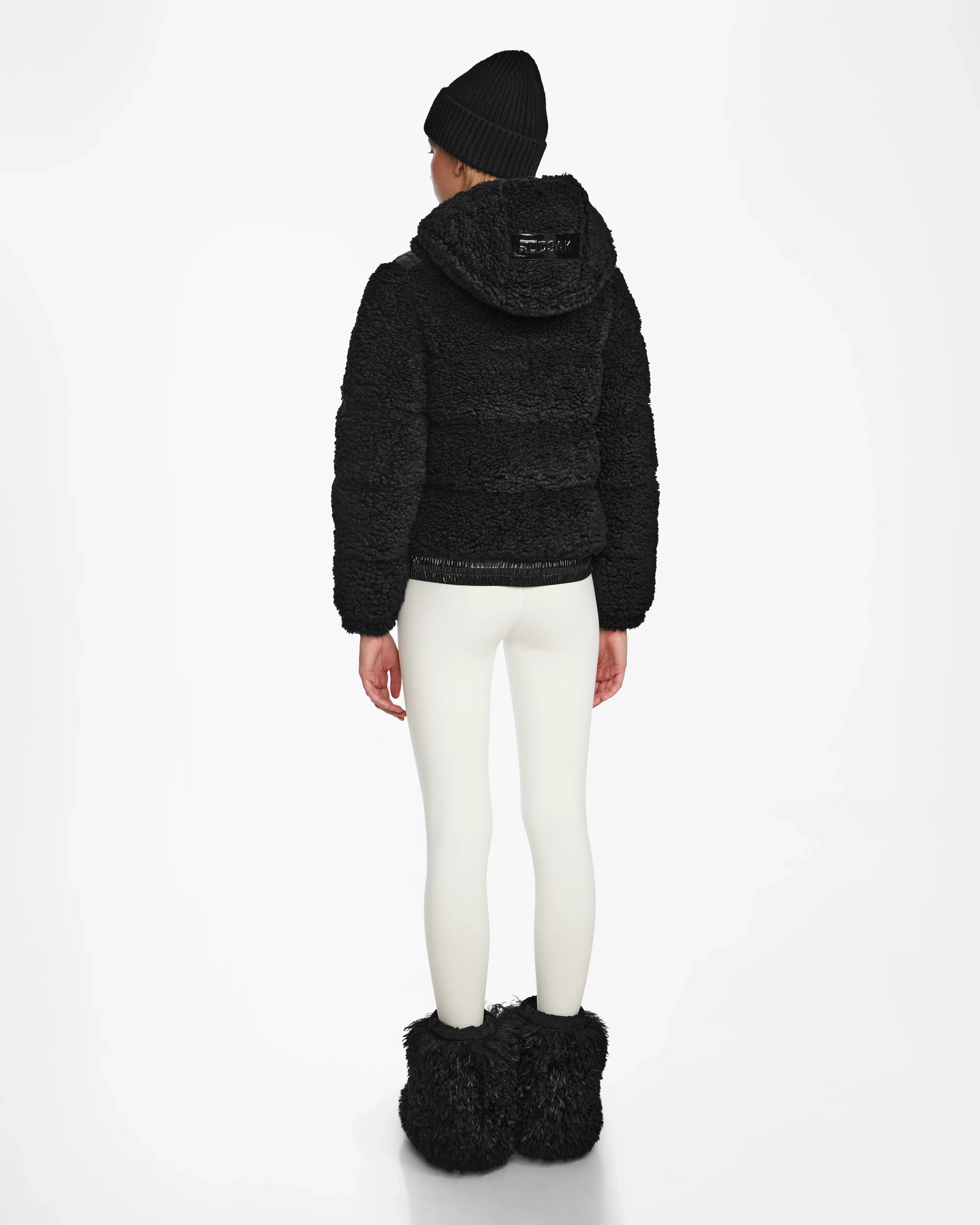 A person faces away, wearing the Rudsak DANI S - 8124537 Black textured cropped ski jacket with hood, paired with white leggings, black fuzzy boots, and a black knit beanie against a plain white background.