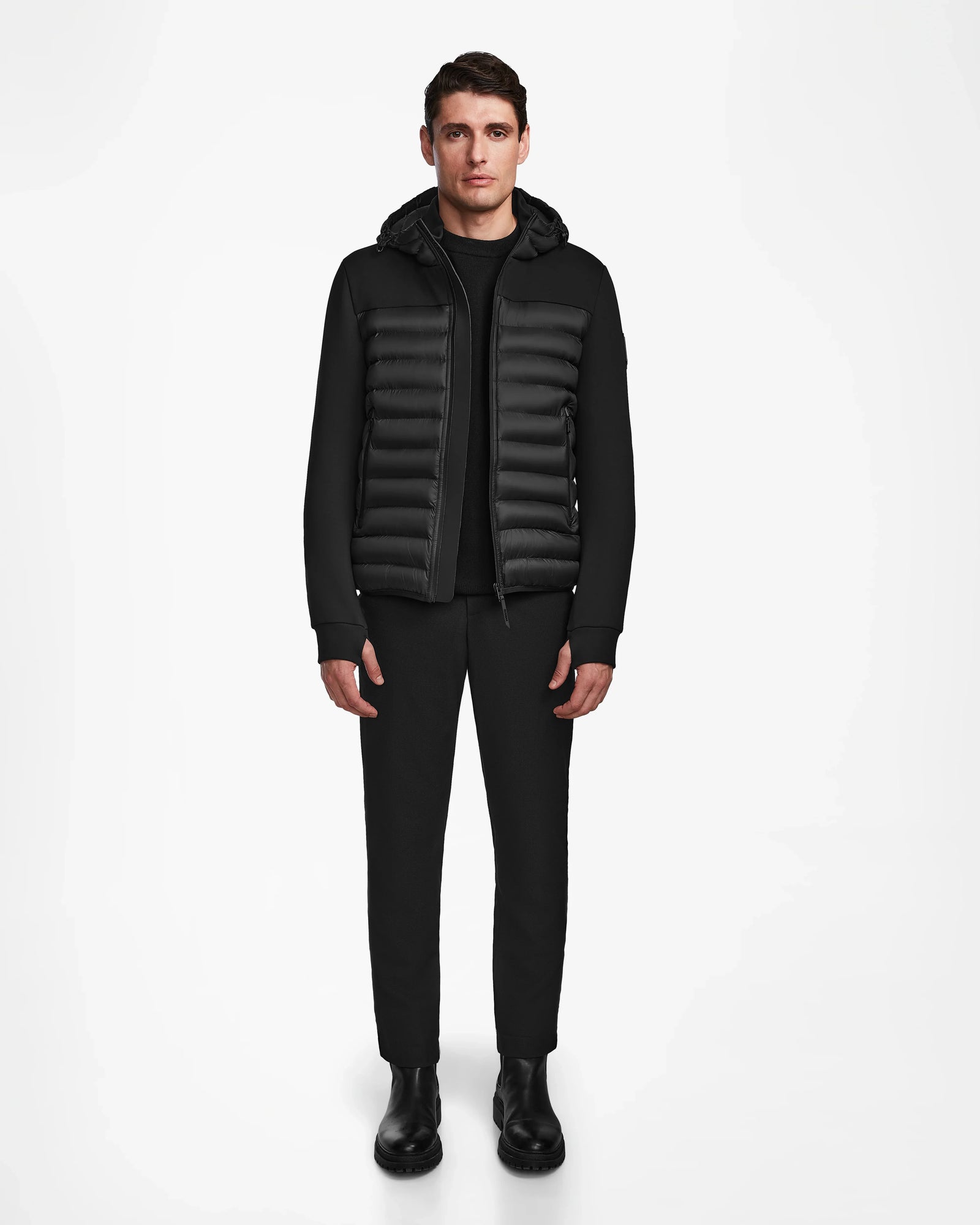 A man faces forward against a white background, wearing Rudsak’s DURIN M - M123515 Black men’s hooded jacket with lightweight down insulation, paired with a black shirt, black pants, and black boots.