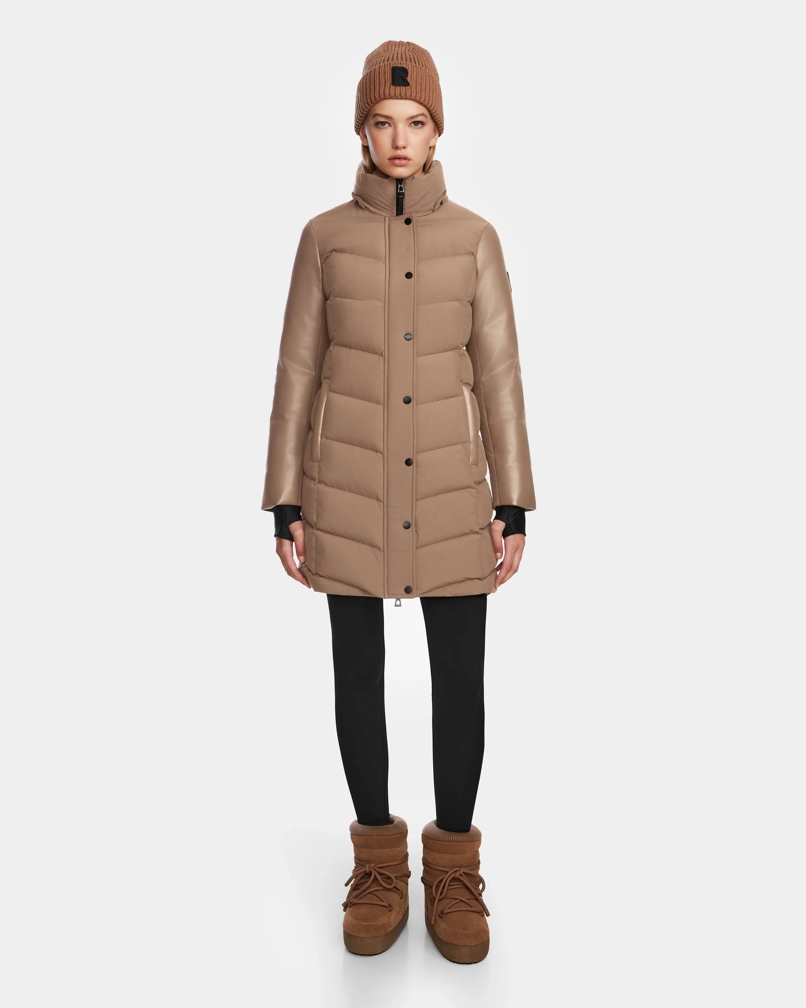 A woman wearing the Rudsak ELSA - L123535 Sand quilted down puffer coat, a matching beanie, black leggings, and brown winter boots stands facing forward against a plain white background.