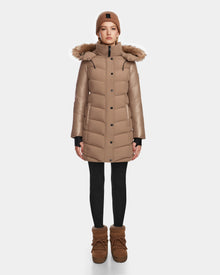 A person stands facing forward, wearing the Rudsak ELSA - L123535 Sand women's quilted down puffer coat with a faux fur-trimmed hood, paired with a tan knit beanie, black leggings, and tan winter boots against a white background.