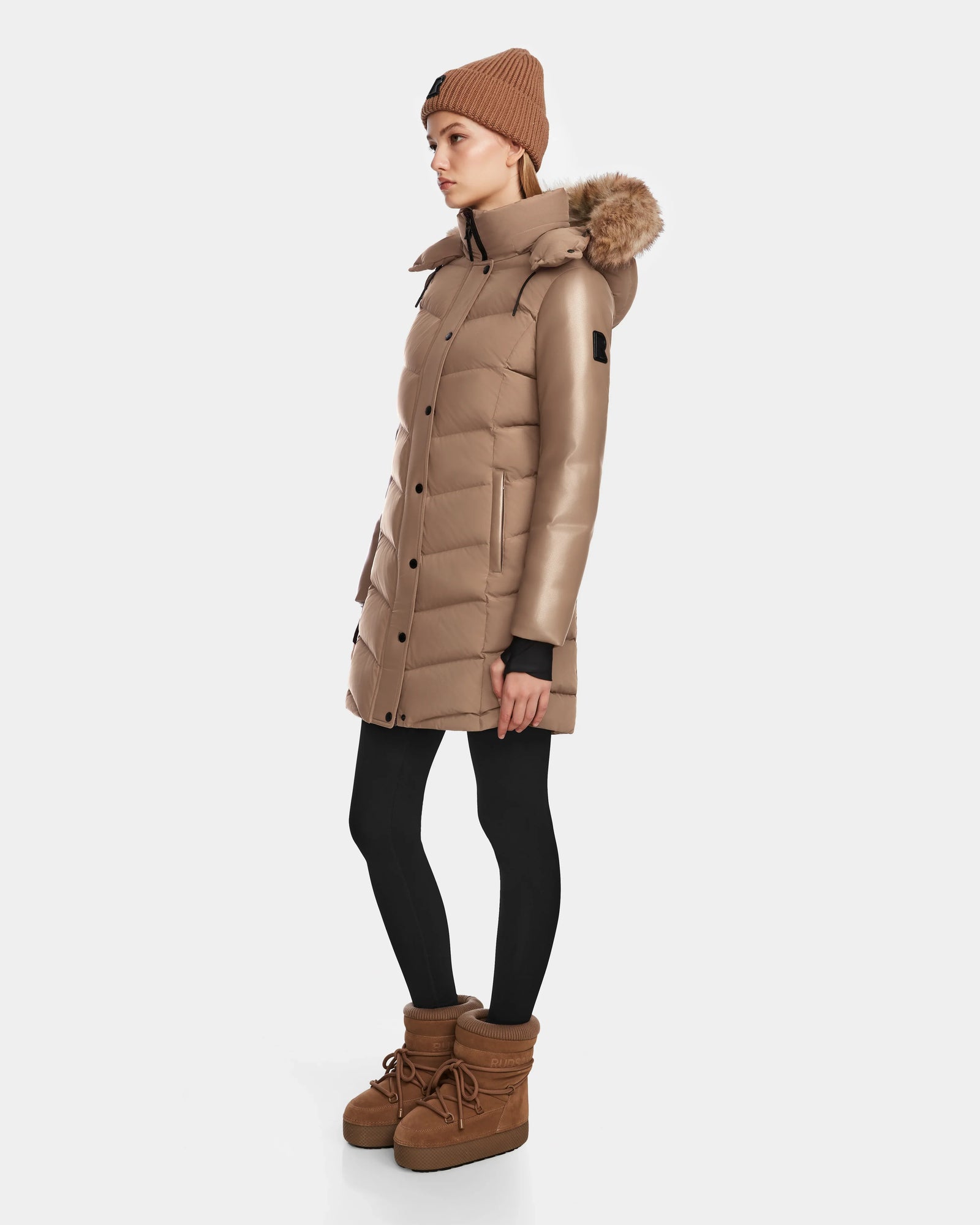 A woman stands sideways against a plain white background, wearing the Rudsak ELSA - L123535 Sand long brown quilted down puffer with a fur-trimmed hood, black leggings, brown winter boots, and a brown knit beanie.