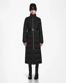 A person stands facing forward against a white background, wearing the Rudsak IZABELLA - L124541 Black women's long down quilted maxi coat, paired with a black beanie, leggings, and boots.