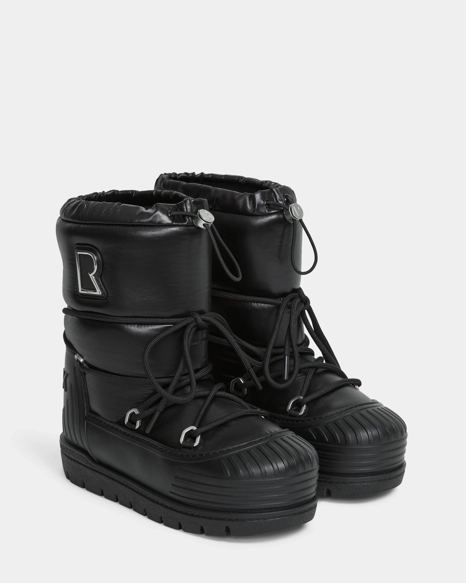 The KIKA - L224506 Black boots by Rudsak are black women’s puffer-style winter boots with chunky rubber soles, drawstring tops, decorative “R” side patches, and ribbed toe and sole detailing.