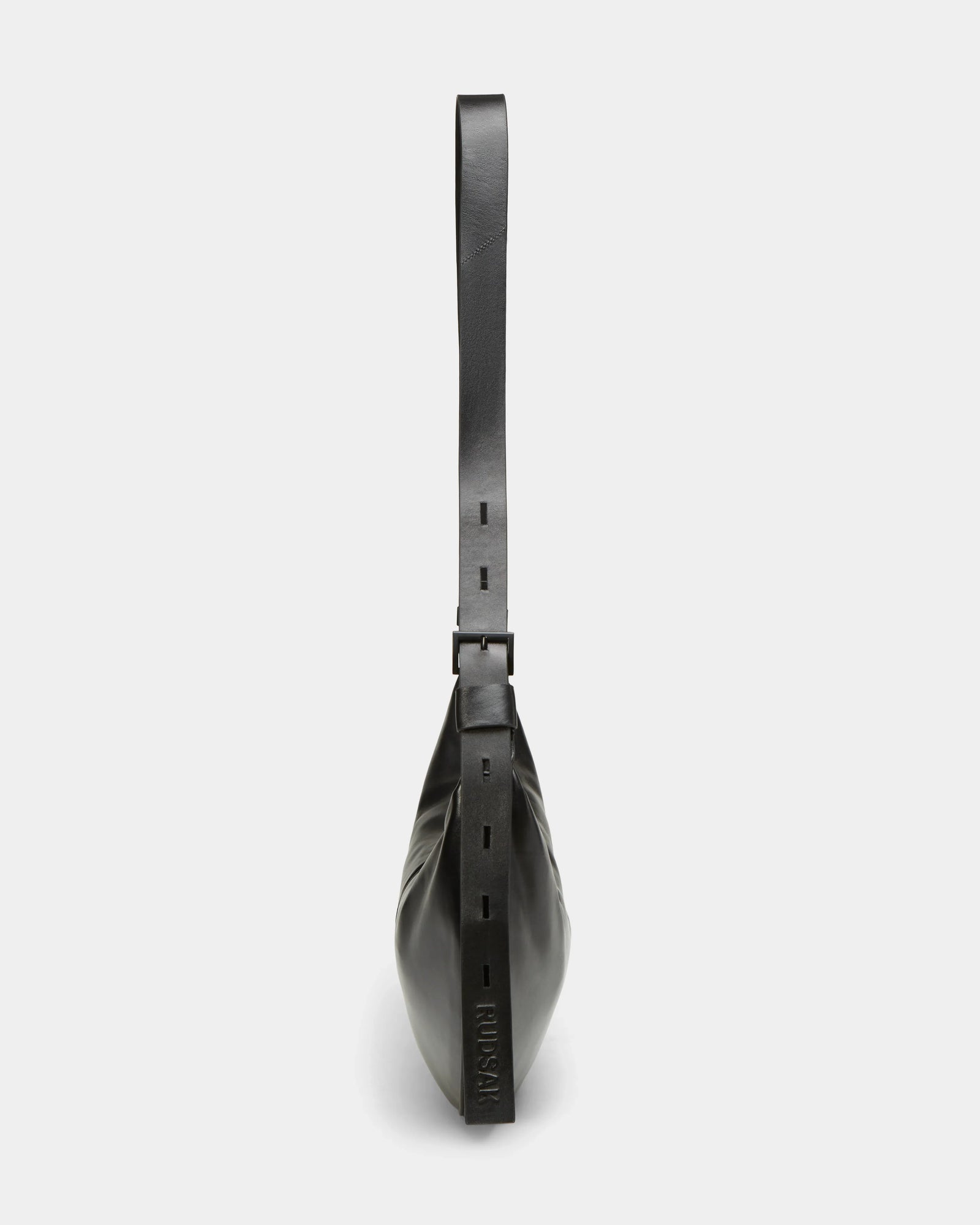 Side view of the Rudsak LANISSA - 8324505 Black leather hobo bag with a wide, adjustable strap and square buckle detail, featuring an embossed logo near the base, shown against a plain white background.