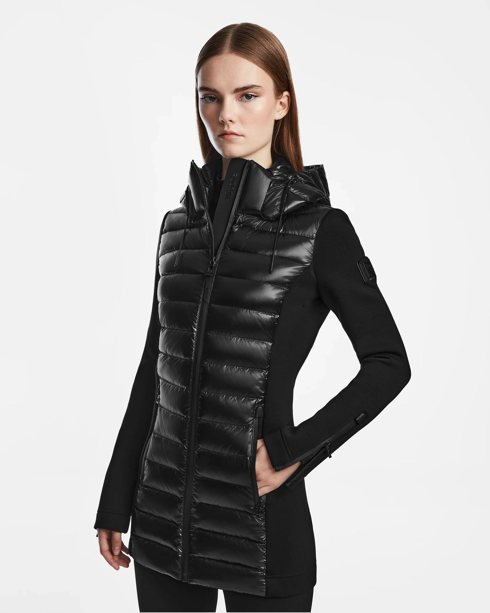 A woman with straight brown hair models the Rudsak MARRY - L121003 BLACK women’s puffer jacket with long sleeves and side pockets, standing against a plain white background.
