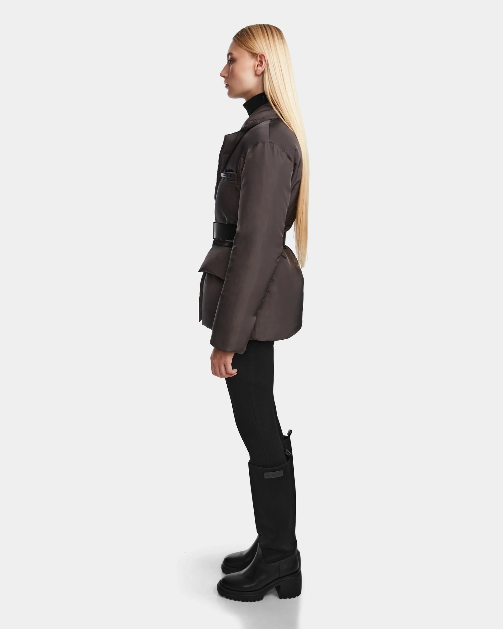 A woman with long blonde hair is seen in profile wearing the Rudsak PAOLA - 8124547 Grey womens down blazer with a belt, styled with black pants and boots against a plain light background.