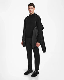 A man stands against a white background wearing a black turtleneck, pants, boots, and draping the Rudsak RICKY R - M123506 Black men's quilted down parka with recycled leather sleeves over his shoulders, looking straight ahead with a neutral expression.