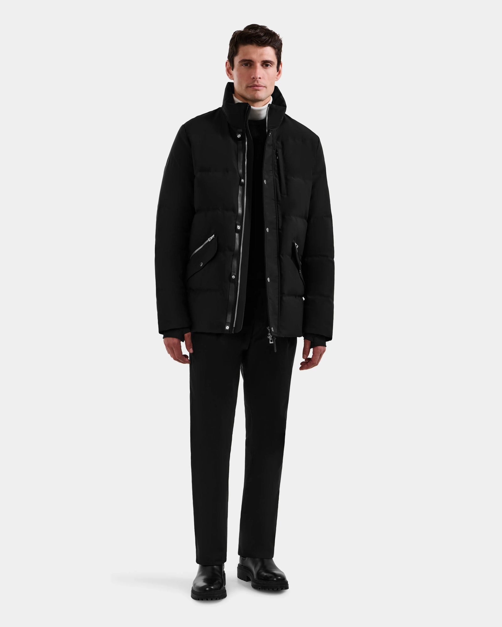 A man stands facing forward against a light gray background, wearing the Rudsak BRADY - M125510 Black men’s quilted down parka with a removable hood, paired with black pants and shoes over a white turtleneck and dark zip-up top.