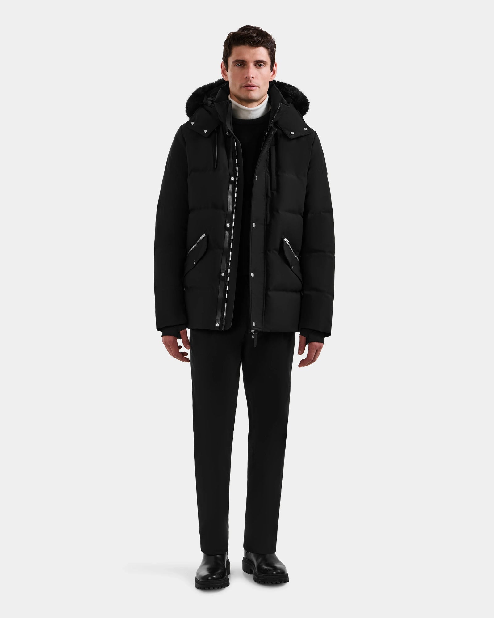 A man faces forward wearing the BRADY - M125510 Black men’s quilted down parka by Rudsak, styled with black pants, black shoes, and a white turtleneck under a black sweater. The background is plain and light-colored.