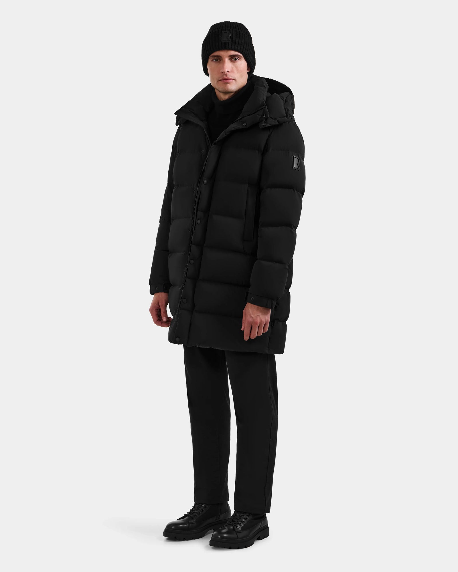 A person showcases winter versatility wearing the Rudsak BRAYDEN M-M - M125535 Black men’s down puffer coat, styled with black pants, a black knit beanie, and black shoes against a plain white background.
