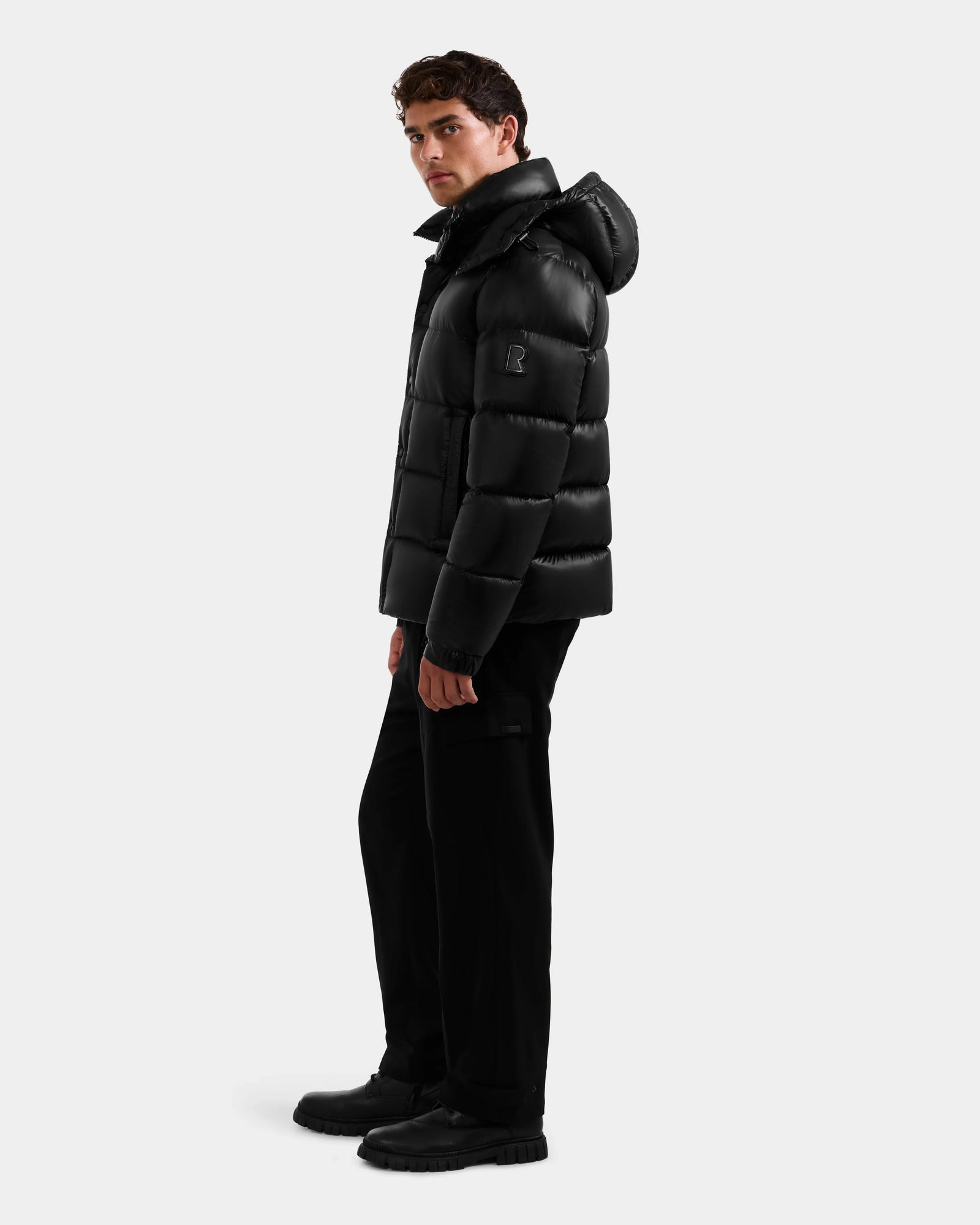 A man stands slightly turned, wearing the Rudsak BRAYDEN S - M125525 Black shiny quilted down puffer jacket with a hood, plus black pants and shoes, against a plain light gray background.