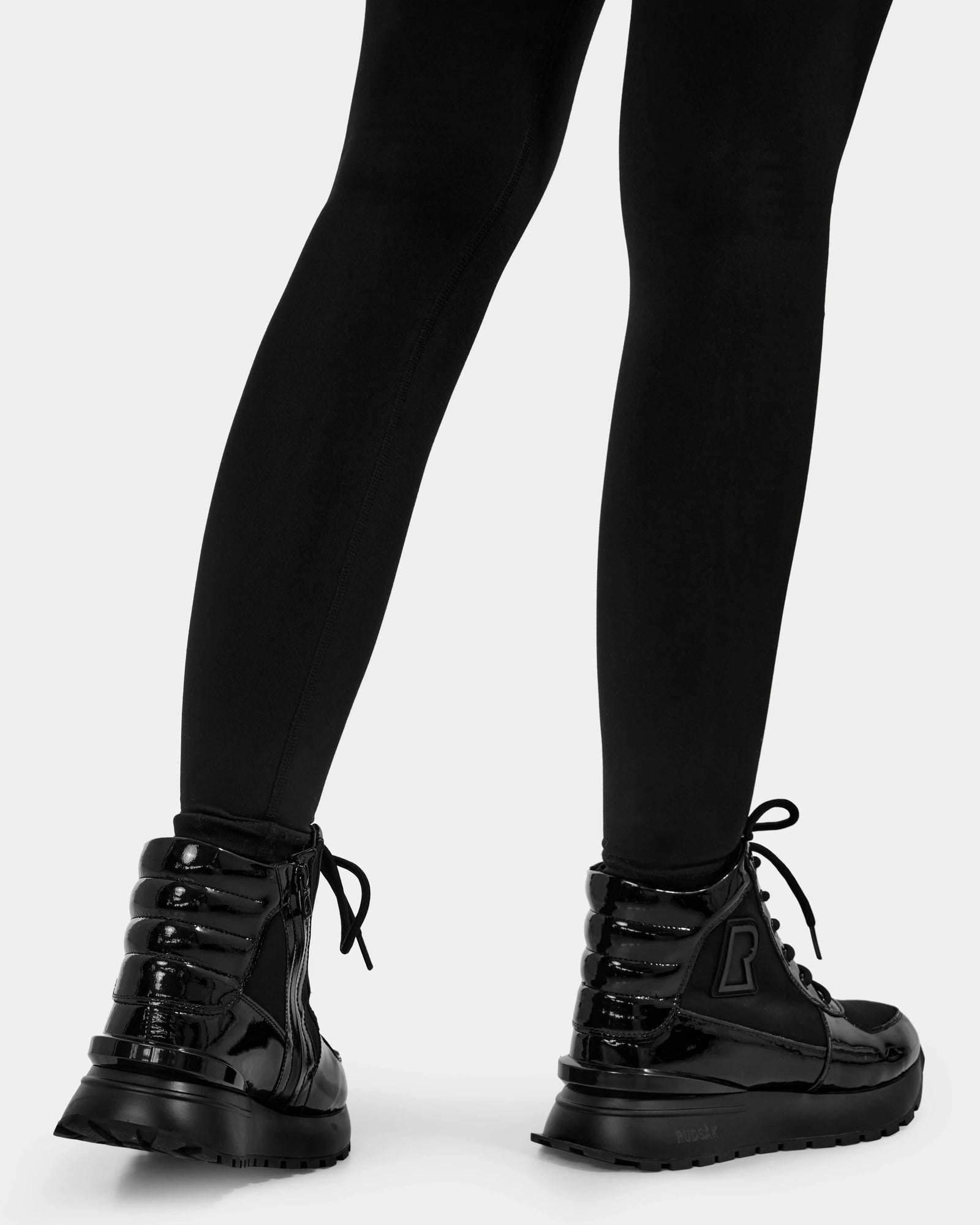 A person wearing black leggings and Rudsak CHELSA - L224505 Black shiny waterproof boots, seen from behind on a white background.