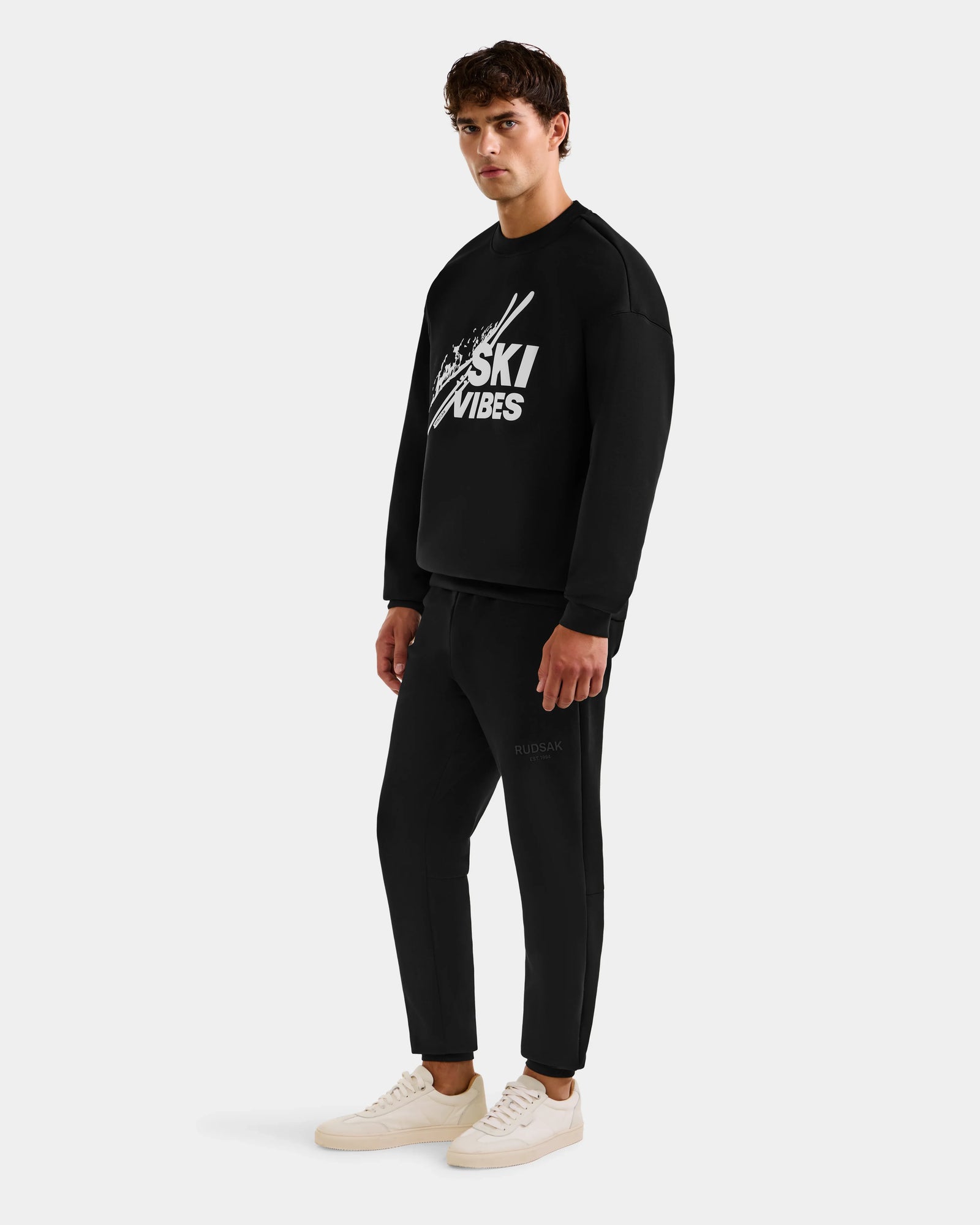 A man wears the Rudsak COLSON - M525501 Black crewneck sweatshirt with the SKI VIBES logo and crossed skis graphic, paired with matching black joggers and white sneakers against a plain white background.