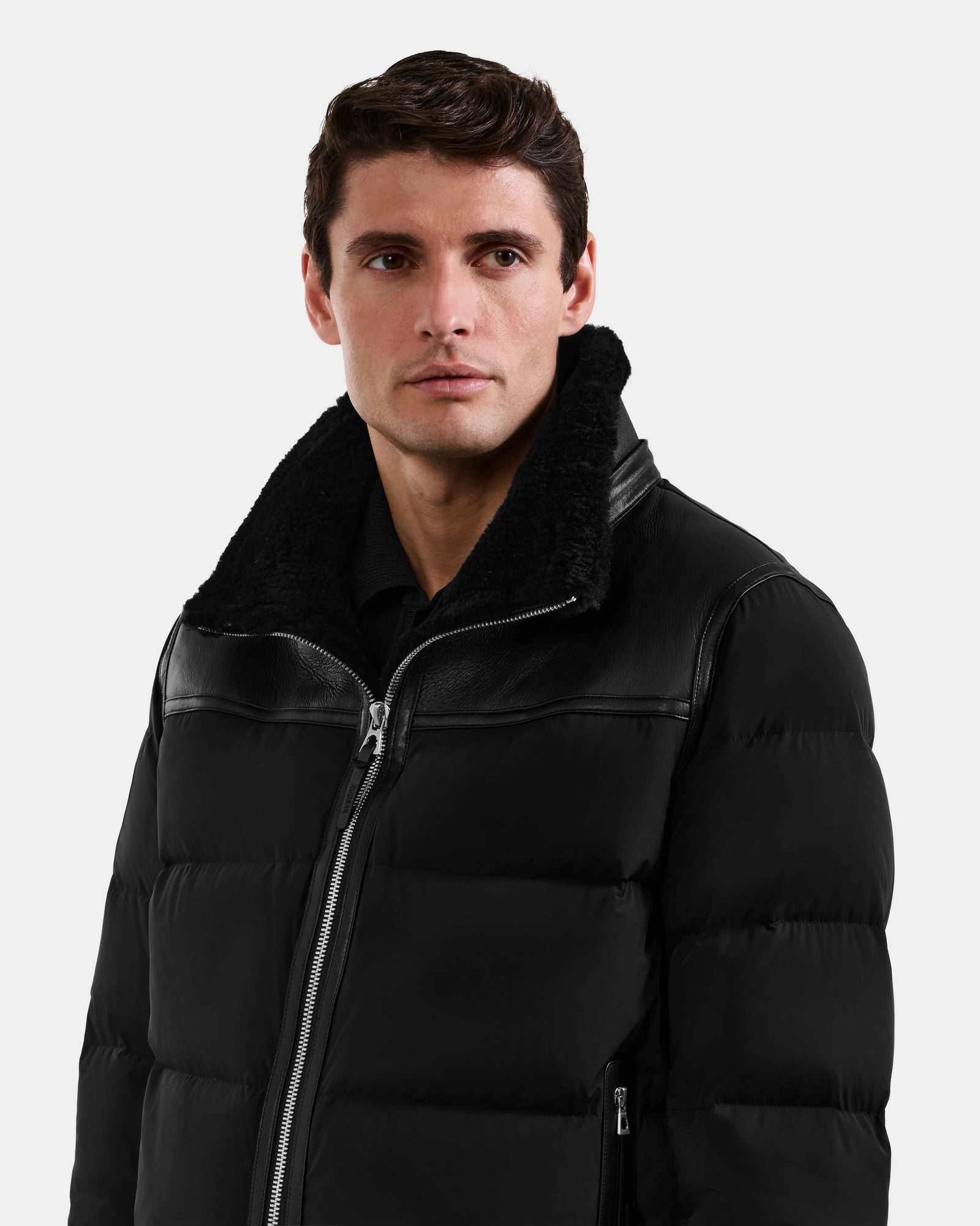 A man with short brown hair wears the Rudsak DAVID - M125508 Black shearling puffer with a fuzzy collar and silver zipper, standing against a plain light background.