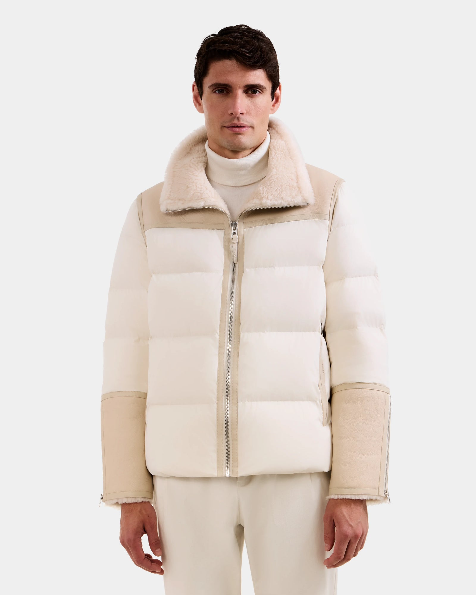 A man wearing the Rudsak DAVID - M125508 Vanilla shearling puffer with a fur-lined collar and light pants stands against a plain light gray background.
