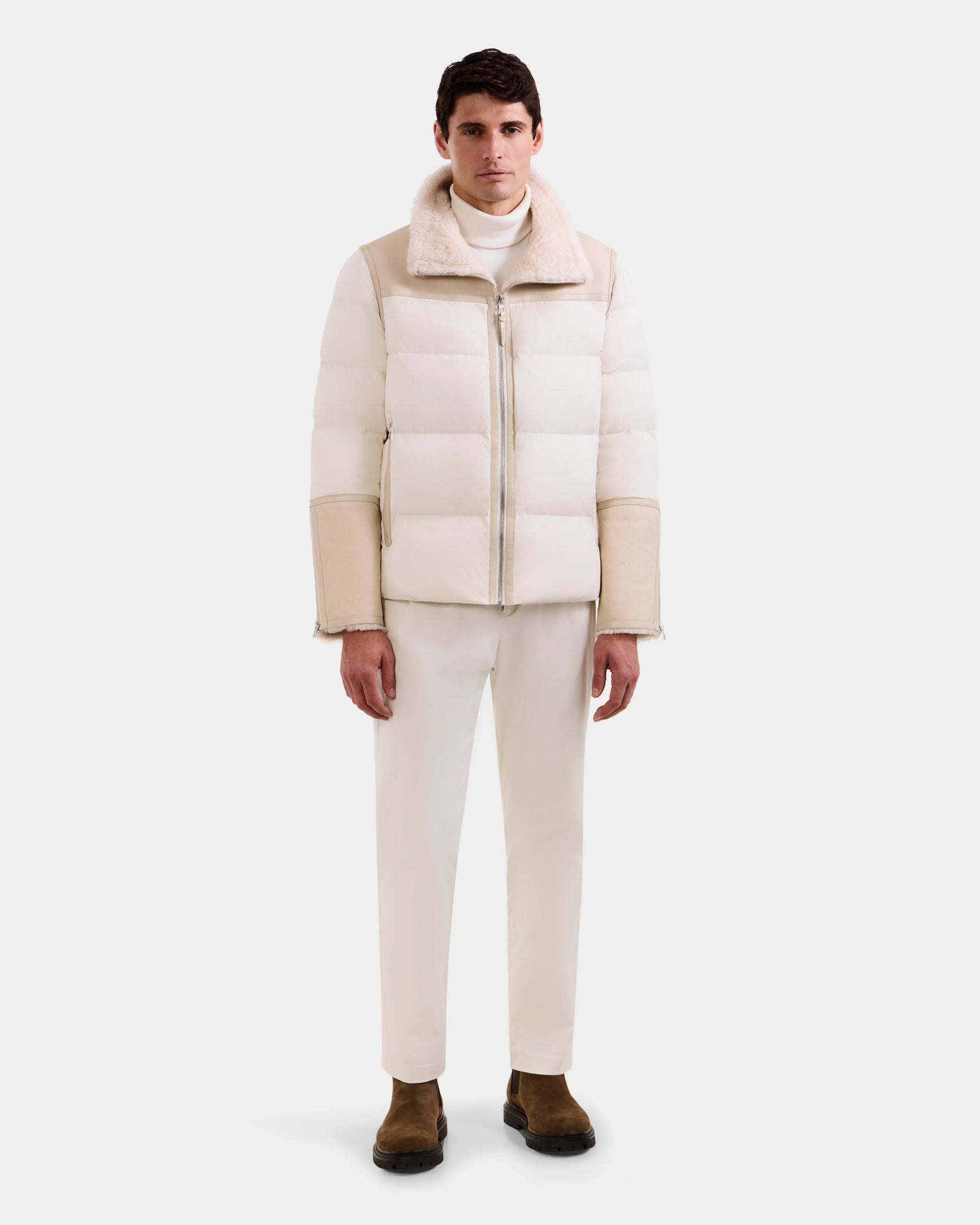 A man faces forward against a plain background, wearing the Rudsak DAVID - M125508 Vanilla men's shearling puffer jacket, white pants, a white turtleneck, and brown boots, with a neutral expression.
