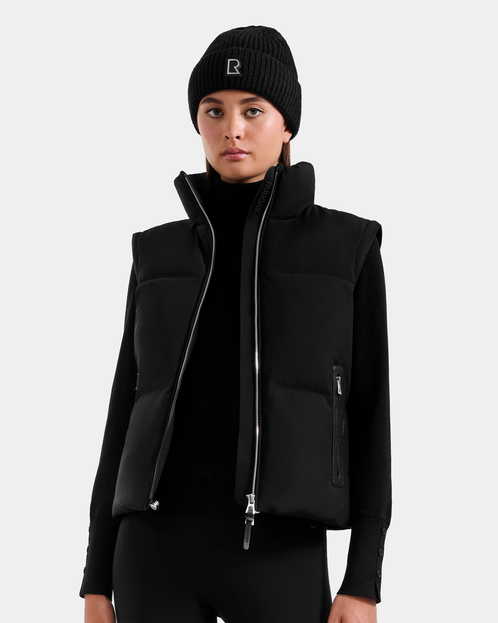 A person wears the Rudsak DELILAH M - L125538 Black women’s down puffer vest with a front zipper, styled with a black beanie, long-sleeve top, and pants, standing against a plain light grey background.