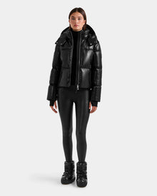 A person faces forward against a plain white background, wearing the Rudsak DELILAH PX - L125537 Black women’s vegan-leather puffer jacket, paired with black leggings and black shoes.
